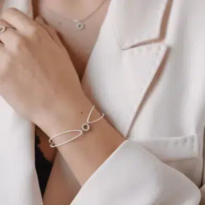 Wrist of a woman wearing a white bracelet with a circle of diamonds in its center. The woman is wearing a beige shirt.